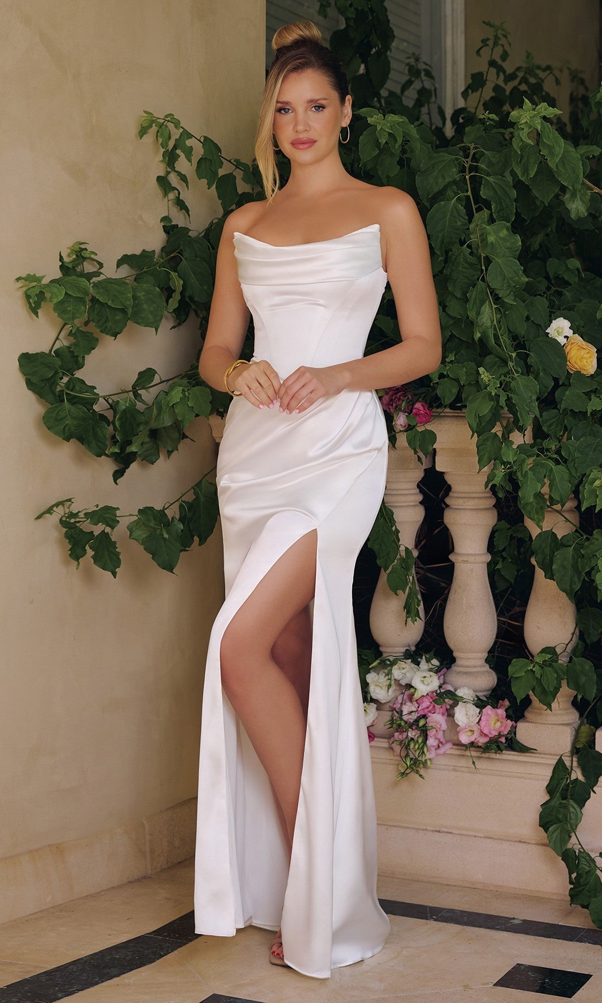 A woman stands before greenery and a stone railing, wearing the Amoris T820W Long Prom Dress in Off White with off-the-shoulder straps and a thigh-high slit.